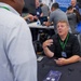 A professional trade show or conference event where attendees are networking and engaging in business discussions. A man in a black shirt with an 'Apex Spaceworks' logo and green lanyard is seated at a booth table, actively conversing with another attendee. The background shows additional booth activity with a sign reading 'Opening Doors for Small Business' and multiple people interacting.