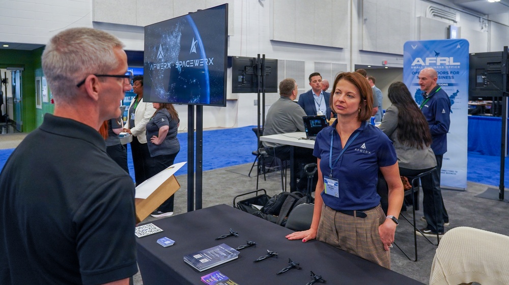 The image shows a trade show or expo booth for AFWERX and SpaceWERX, military innovation programs associated with the U.S. Air Force. A woman wearing an AFWERX polo shirt is engaged in conversation with a man at the booth table. In the background, additional attendees and staff are visible near an AFRL (Air Force Research Laboratory) Small Business Office banner.