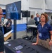 The image shows a trade show or expo booth for AFWERX and SpaceWERX, military innovation programs associated with the U.S. Air Force. A woman wearing an AFWERX polo shirt is engaged in conversation with a man at the booth table. In the background, additional attendees and staff are visible near an AFRL (Air Force Research Laboratory) Small Business Office banner.
