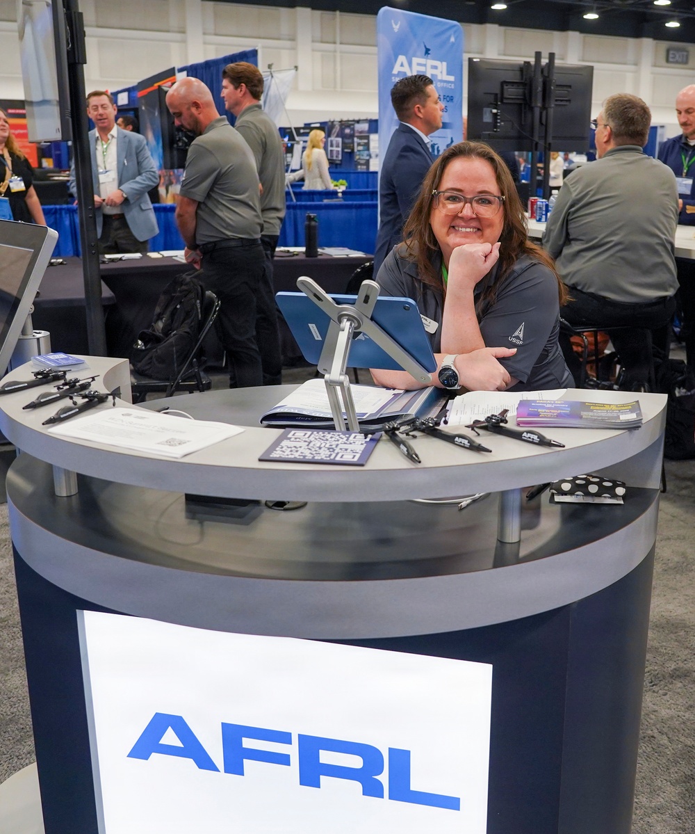 A woman wearing a USSF (United States Space Force) branded polo shirt is staffing an AFRL (Air Force Research Laboratory) exhibition booth at what appears to be a professional conference or trade show. She is smiling and posing for the camera while leaning on the circular reception desk. The booth features branded materials, small aircraft models, a tablet on a stand, and a QR code display.