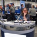 A woman wearing a USSF (United States Space Force) branded polo shirt is staffing an AFRL (Air Force Research Laboratory) exhibition booth at what appears to be a professional conference or trade show. She is smiling and posing for the camera while leaning on the circular reception desk. The booth features branded materials, small aircraft models, a tablet on a stand, and a QR code display.