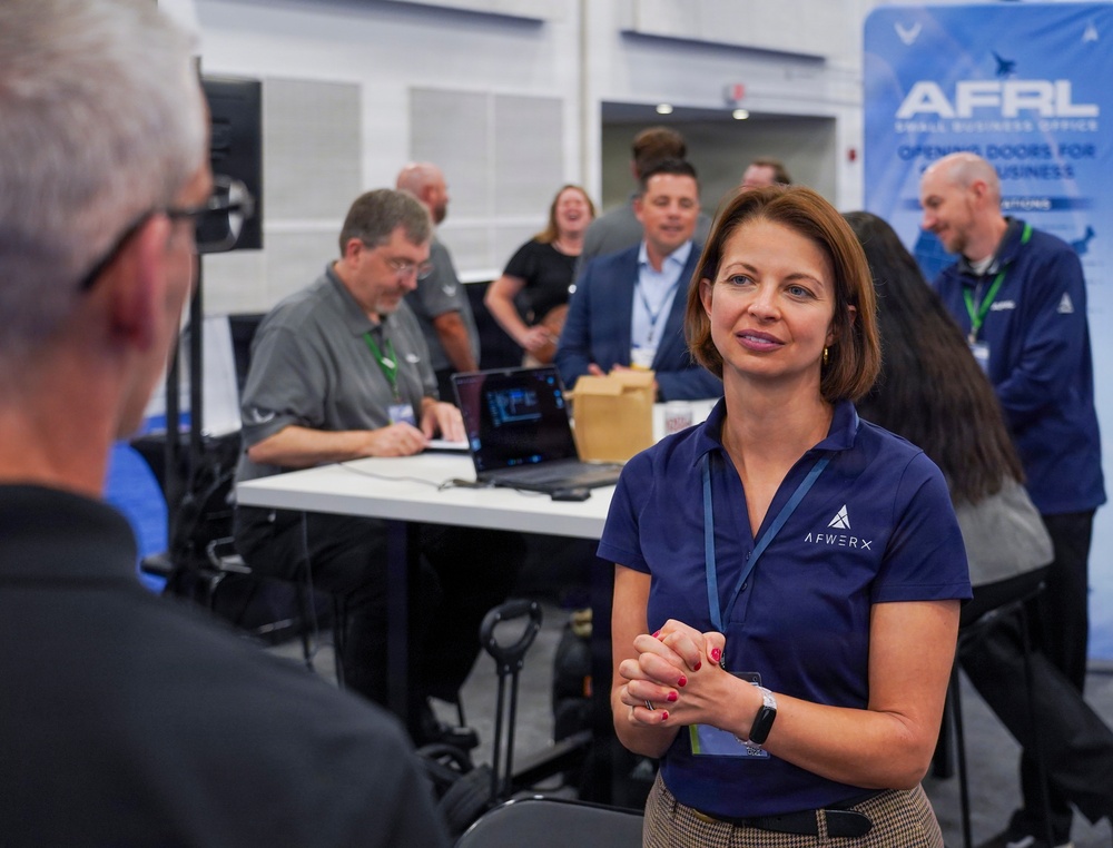 A professional event or conference setting featuring an AFWERX and AFRL Small Business Office booth or meeting area. A woman wearing a navy blue AFWERX polo shirt is engaged in conversation with another person in the foreground. Multiple attendees are visible in the background, some seated at tables with laptops, others standing and conversing.