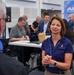 A professional event or conference setting featuring an AFWERX and AFRL Small Business Office booth or meeting area. A woman wearing a navy blue AFWERX polo shirt is engaged in conversation with another person in the foreground. Multiple attendees are visible in the background, some seated at tables with laptops, others standing and conversing.