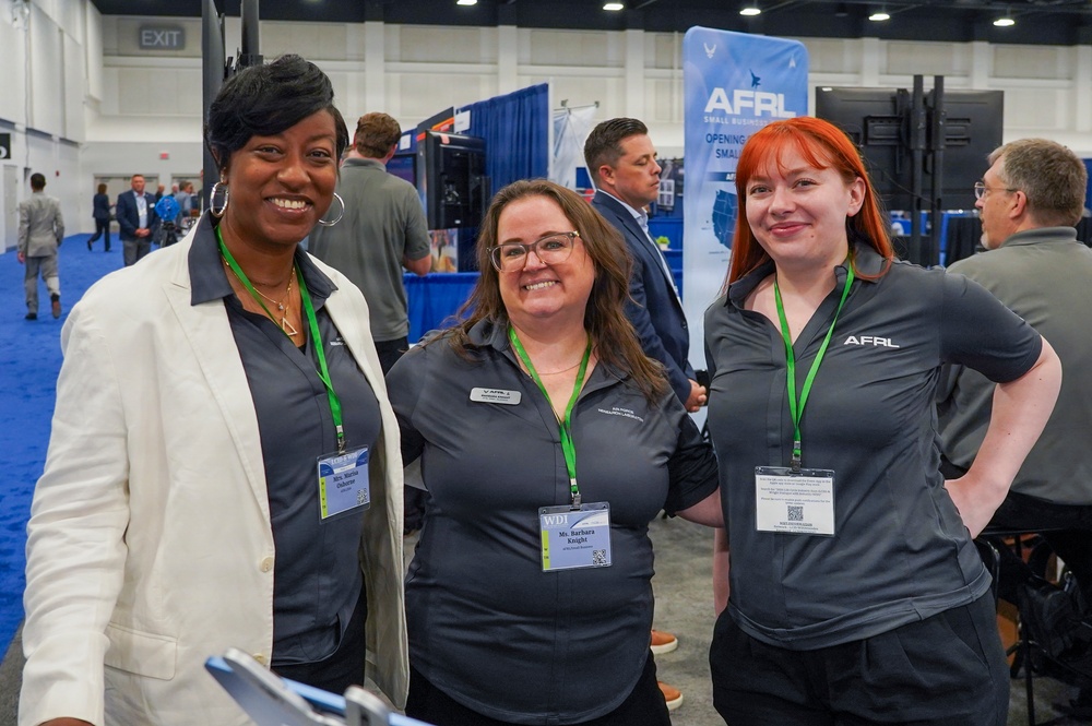 Three women pose together at what appears to be a professional conference or trade show, with an AFRL (Air Force Research Laboratory) booth visible in the background. Two of the women are wearing dark gray AFRL-branded polo shirts with green lanyards and name badges, while the third wears a white blazer over a dark shirt. The event appears to be a small business or defense industry expo.