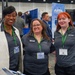 Three women pose together at what appears to be a professional conference or trade show, with an AFRL (Air Force Research Laboratory) booth visible in the background. Two of the women are wearing dark gray AFRL-branded polo shirts with green lanyards and name badges, while the third wears a white blazer over a dark shirt. The event appears to be a small business or defense industry expo.