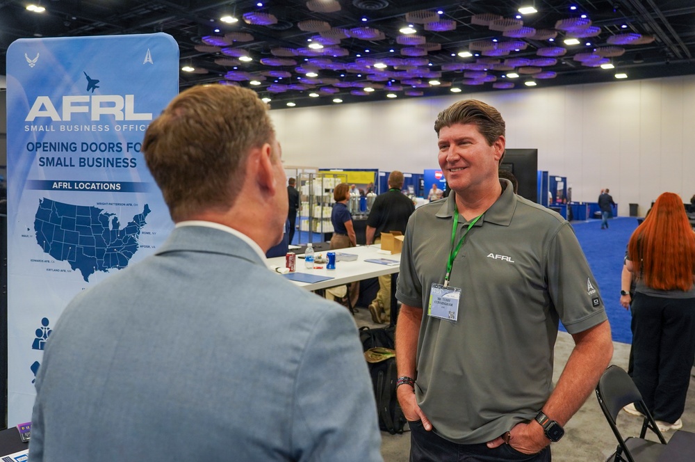 The image shows a professional conference or trade show event featuring an AFRL (Air Force Research Laboratory) Small Business Office booth. Two men are engaged in conversation near the booth's banner, which reads 'Opening Doors for Small Business.' The setting appears to be a large convention center with multiple exhibitor booths visible in the background.