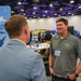 The image shows a professional conference or trade show event featuring an AFRL (Air Force Research Laboratory) Small Business Office booth. Two men are engaged in conversation near the booth's banner, which reads 'Opening Doors for Small Business.' The setting appears to be a large convention center with multiple exhibitor booths visible in the background.