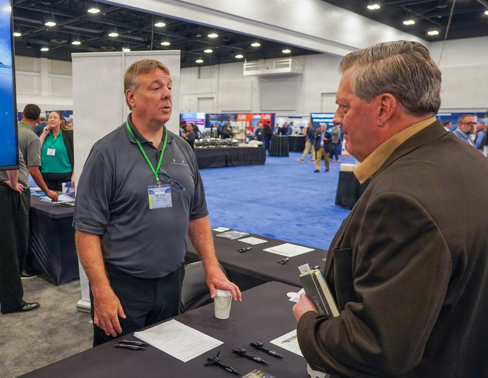 Two men are engaged in conversation at a trade show or conference exhibition booth. The man on the left is wearing a gray polo shirt with a green lanyard and name badge, appearing to be a vendor or exhibitor. The setting is a large convention center floor with multiple vendor booths visible in the background.