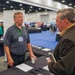 Two men are engaged in conversation at a trade show or conference exhibition booth. The man on the left is wearing a gray polo shirt with a green lanyard and name badge, appearing to be a vendor or exhibitor. The setting is a large convention center floor with multiple vendor booths visible in the background.
