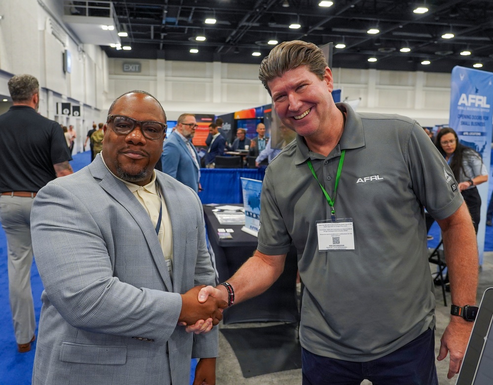 Two men are shaking hands at what appears to be a professional trade show or conference. The man on the right is wearing an AFRL (Air Force Research Laboratory) branded polo shirt with a green lanyard and name badge. The event appears to be a business or defense industry expo with multiple vendor booths visible in the background.