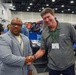Two men are shaking hands at what appears to be a professional trade show or conference. The man on the right is wearing an AFRL (Air Force Research Laboratory) branded polo shirt with a green lanyard and name badge. The event appears to be a business or defense industry expo with multiple vendor booths visible in the background.