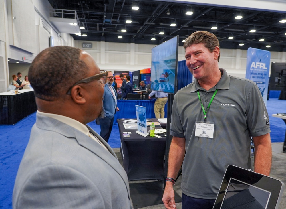 Two men are engaged in a friendly conversation at what appears to be a professional trade show or conference expo floor. The man on the right is wearing an AFRL (Air Force Research Laboratory) branded polo shirt and green lanyard badge, suggesting he is a representative at the AFRL booth. The setting features multiple vendor booths, blue carpeting, and conference center infrastructure.