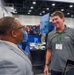 Two men are engaged in a friendly conversation at what appears to be a professional trade show or conference expo floor. The man on the right is wearing an AFRL (Air Force Research Laboratory) branded polo shirt and green lanyard badge, suggesting he is a representative at the AFRL booth. The setting features multiple vendor booths, blue carpeting, and conference center infrastructure.