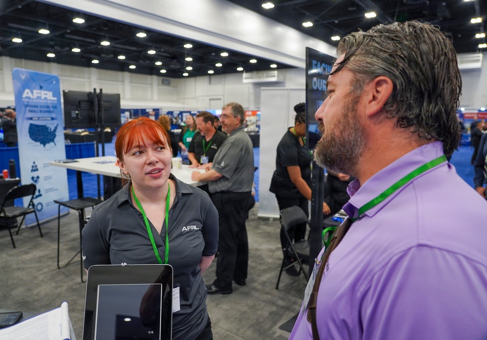 The image shows a trade show or expo floor where representatives from AFRL (Air Force Research Laboratory) are engaging with attendees. A young woman with red hair wearing an AFRL-branded polo shirt and green lanyard is speaking with a bearded man in a purple shirt who also wears a green lanyard. Multiple exhibitor booths and other attendees are visible in the background.