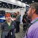 The image shows a trade show or expo floor where representatives from AFRL (Air Force Research Laboratory) are engaging with attendees. A young woman with red hair wearing an AFRL-branded polo shirt and green lanyard is speaking with a bearded man in a purple shirt who also wears a green lanyard. Multiple exhibitor booths and other attendees are visible in the background.