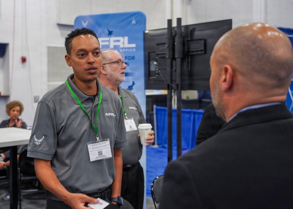 The image shows a professional event or conference where representatives from AFRL (Air Force Research Laboratory) are engaging with attendees. Two men wearing gray AFRL polo shirts with U.S. Air Force insignia and green lanyards are conversing with a visitor in a black jacket. The setting appears to be an exhibition hall or trade show booth.