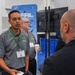 The image shows a professional event or conference where representatives from AFRL (Air Force Research Laboratory) are engaging with attendees. Two men wearing gray AFRL polo shirts with U.S. Air Force insignia and green lanyards are conversing with a visitor in a black jacket. The setting appears to be an exhibition hall or trade show booth.