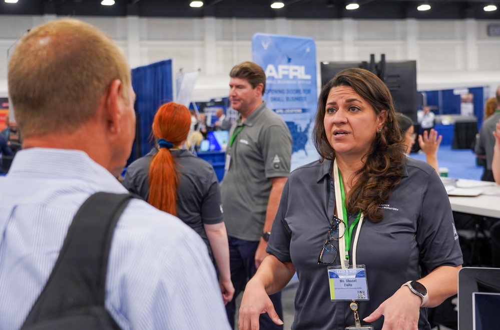The image shows a professional conference or trade show environment featuring an AFRL (Air Force Research Laboratory) booth. A woman wearing a gray polo shirt and a name badge is engaged in conversation with a man whose back is to the camera. Multiple staff members in matching gray shirts are visible throughout the booth area.