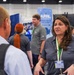 The image shows a professional conference or trade show environment featuring an AFRL (Air Force Research Laboratory) booth. A woman wearing a gray polo shirt and a name badge is engaged in conversation with a man whose back is to the camera. Multiple staff members in matching gray shirts are visible throughout the booth area.