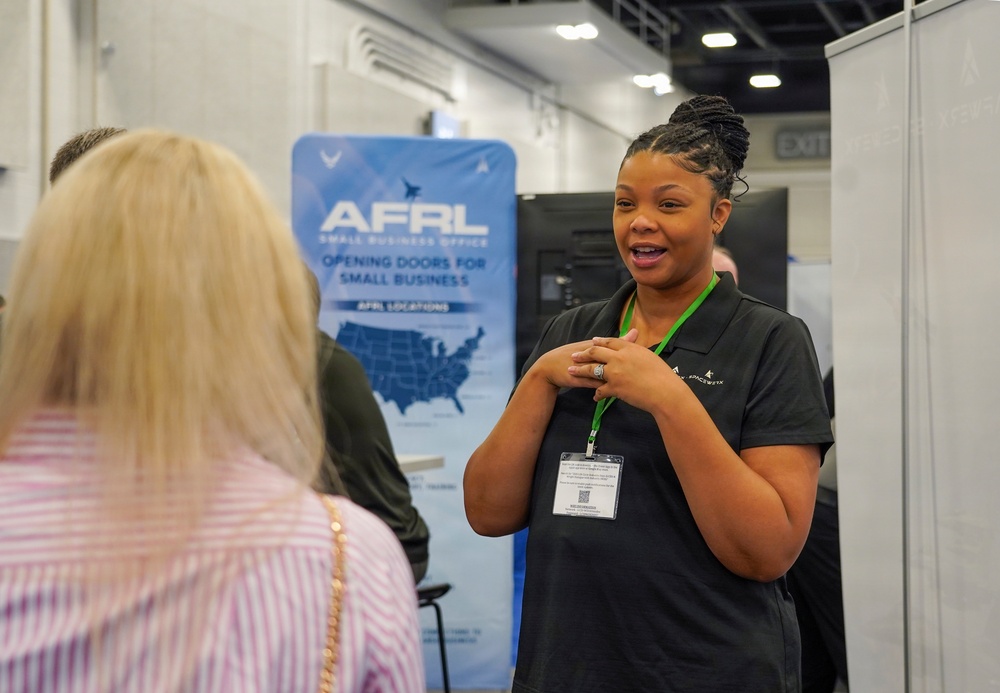A woman wearing a dark polo shirt and green lanyard with a badge is speaking to another person at what appears to be a professional conference or trade show. Behind her is an AFRL (Air Force Research Laboratory) Small Business Office banner reading 'Opening Doors for Small Business.' The setting is an indoor exhibition hall with booth displays and overhead lighting.