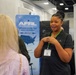 A woman wearing a dark polo shirt and green lanyard with a badge is speaking to another person at what appears to be a professional conference or trade show. Behind her is an AFRL (Air Force Research Laboratory) Small Business Office banner reading 'Opening Doors for Small Business.' The setting is an indoor exhibition hall with booth displays and overhead lighting.