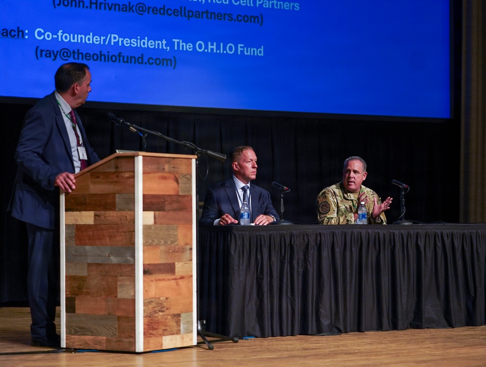 A panel discussion or conference event taking place on a stage, featuring three men — one standing at a wooden podium and two seated at a draped table. A large blue presentation screen in the background displays contact information including references to 'Red Cell Partners' and 'The O.H.I.O Fund.' One of the seated individuals is wearing military camouflage uniform while the others are in civilian business attire.