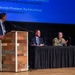 A panel discussion or conference event taking place on a stage, featuring three men — one standing at a wooden podium and two seated at a draped table. A large blue presentation screen in the background displays contact information including references to 'Red Cell Partners' and 'The O.H.I.O Fund.' One of the seated individuals is wearing military camouflage uniform while the others are in civilian business attire.