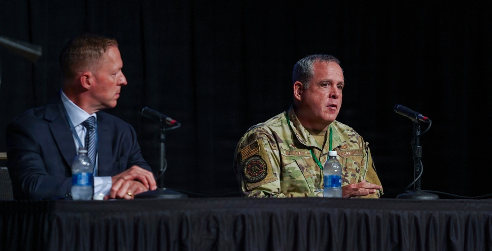 Two individuals are seated at a draped panel table during what appears to be a formal conference or briefing. One person is wearing a military uniform in MultiCam/OCP pattern with visible patches, while the other is dressed in civilian business attire. The military individual appears to be speaking, with microphones positioned in front of both participants.