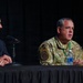 Two individuals are seated at a draped panel table during what appears to be a formal conference or briefing. One person is wearing a military uniform in MultiCam/OCP pattern with visible patches, while the other is dressed in civilian business attire. The military individual appears to be speaking, with microphones positioned in front of both participants.