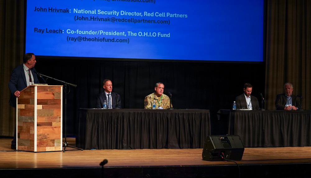 A panel discussion or conference event is taking place on a stage, with four panelists seated at a draped table and a moderator standing at a wooden podium. A large blue projection screen behind them displays speaker names and affiliations, including John Hrivnak (National Security Director, Red Cell Partners) and Ray Leach (Co-founder/President, The O.H.I.O Fund). One panelist is wearing military camouflage attire, while the others are in civilian business dress.