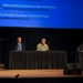 A panel discussion or conference event is taking place on a stage, with four panelists seated at a draped table and a moderator standing at a wooden podium. A large blue projection screen behind them displays speaker names and affiliations, including John Hrivnak (National Security Director, Red Cell Partners) and Ray Leach (Co-founder/President, The O.H.I.O Fund). One panelist is wearing military camouflage attire, while the others are in civilian business dress.