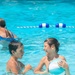 Two children are playing in an outdoor swimming pool on a sunny day. One child is holding a teal and white beach ball while laughing, and the other child faces them in an interactive, playful manner. A third person is partially visible in the background near a lane divider rope.