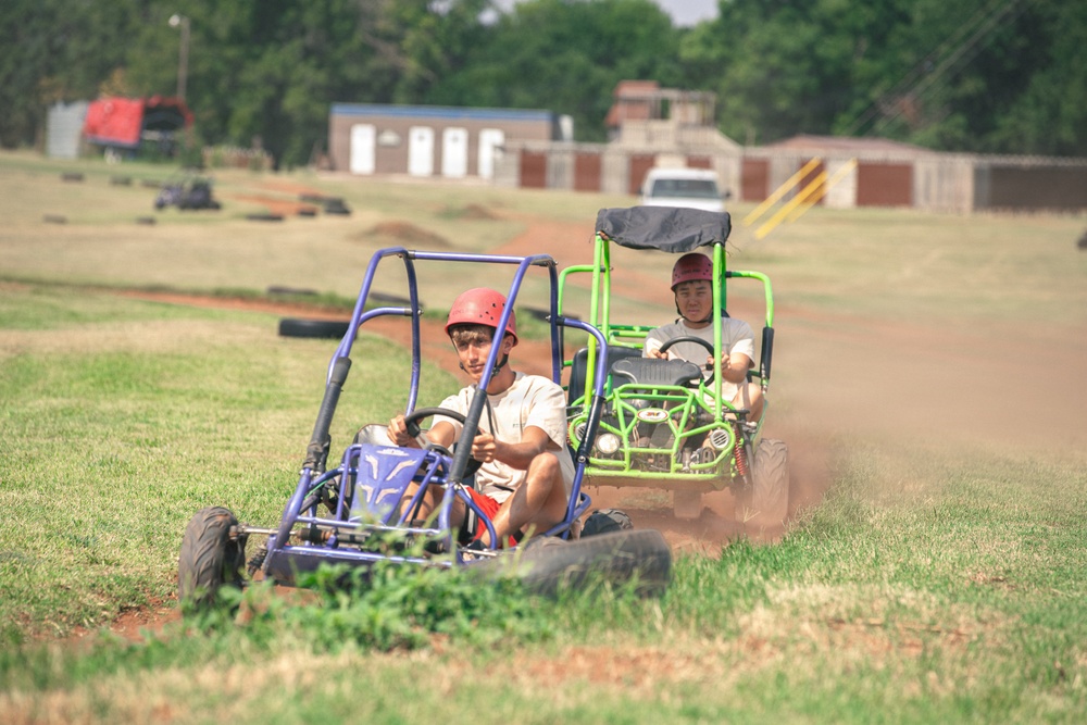 Two young people are riding off-road go-karts on a dirt track surrounded by grass. The foreground rider operates a blue go-kart while wearing a red helmet, and a second rider follows closely in a green go-kart also wearing a red cap. The background shows buildings, trees, and what appears to be a recreational go-kart facility.