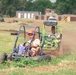 Two young people are riding off-road go-karts on a dirt track surrounded by grass. The foreground rider operates a blue go-kart while wearing a red helmet, and a second rider follows closely in a green go-kart also wearing a red cap. The background shows buildings, trees, and what appears to be a recreational go-kart facility.