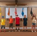Seven children and young teens stand in a line inside what appears to be a camp or church hall, each holding a tall wooden flagpole with a colored pennant flag. The flags vary in color including light blue, yellow, orange, red, blue, black, and olive green, each appearing to bear a distinct emblem or logo. A large wooden cross is visible on the wall behind them, along with a projection screen, suggesting a religious camp or youth organization setting.