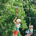 Two children are riding parallel ziplines outdoors, suspended in the air with safety harnesses and helmets. The foreground child is smiling and wearing red shorts with a white Petzl helmet, while a second child is visible in the background on a separate line. The setting appears to be a summer camp or adventure park with large green trees in the background.