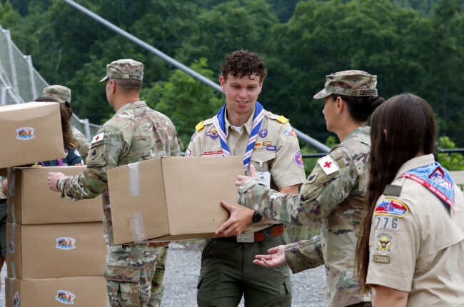 Military personnel and Boy Scouts are collaborating to unload and pass cardboard boxes in an outdoor setting surrounded by green trees. The boxes are marked with stickers, and participants include both uniformed soldiers in camouflage and scouts in tan uniforms with merit badges and neckerchiefs. The scene appears to be a supply distribution or humanitarian aid effort at what may be a scout camp or training facility.