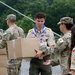 Military personnel and Boy Scouts are collaborating to unload and pass cardboard boxes in an outdoor setting surrounded by green trees. The boxes are marked with stickers, and participants include both uniformed soldiers in camouflage and scouts in tan uniforms with merit badges and neckerchiefs. The scene appears to be a supply distribution or humanitarian aid effort at what may be a scout camp or training facility.
