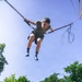 A young boy is suspended in mid-air while using a bungee trampoline attraction, harnessed with safety straps and bungee cords attached to a metal frame above. The photo is taken from a low angle looking upward, capturing the boy against a clear blue sky with green trees in the background. He appears excited and is gripping the bungee cords with both hands while his legs are bent mid-bounce.