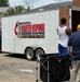 A white trailer belonging to United Methodists Disaster Response Ministries, WV Annual Conference, is parked at a brick building while several people unload or load supplies. The trailer bears the text 'Partners of WV VOAD' (Voluntary Organizations Active in Disaster). This appears to be a disaster relief supply operation with volunteers working at the trailer's open rear doors.
