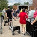 A group of volunteers or workers are unloading supplies from an open trailer truck outside a brick building, likely during a community relief or donation effort. Multiple people are using hand trucks, shopping carts, and utility carts to transport boxes and pallets of bottled water. The scene suggests an organized distribution or collection event taking place outdoors in warm weather.