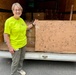 A smiling older woman wearing a bright yellow/neon green t-shirt stands beside a large cardboard panel covered in hand-drawn doodles and motivational messages. She is positioned at the open rear of a cargo trailer or moving truck loaded with cardboard boxes. The cardboard panel appears to be a community art piece with drawings, signatures, and encouraging phrases.