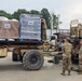 Military personnel in camouflage uniforms are loading or securing pallets of supplies onto a military flatbed trailer. The supplies, including cases of bottled water and boxed goods, are labeled with 'Scouting America' branding, suggesting a humanitarian or disaster relief operation. Several soldiers are coordinating the logistics while a forklift-type military vehicle is visible on the right.