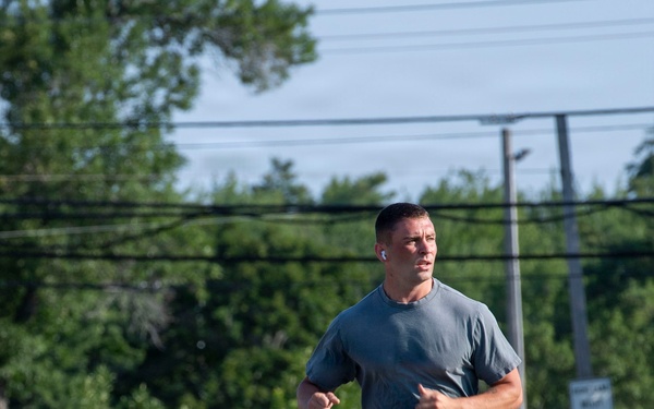A male runner is captured mid-stride on a gravel path, wearing athletic clothing including a gray t-shirt, olive shorts, black socks, and black running shoes. He has a wireless earbud in his right ear and appears to be running with focused intensity. The setting appears to be an outdoor recreational or military installation area with chain-link fencing, utility poles, and green trees in the background.