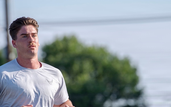 A young adult male is jogging outdoors wearing a light gray U.S. Air Force physical training t-shirt and navy blue shorts. The image is captured with a shallow depth of field, keeping the subject sharp while the background of trees, utility poles, and sky is blurred. The lighting suggests early morning or late afternoon conditions.