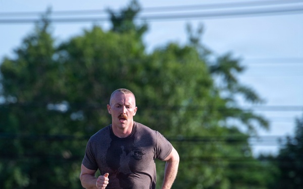 A muscular man with a mustache is running at pace along a gravel path or road. He is wearing a dark gray t-shirt with a graphic, black running shorts, and white athletic shoes, with a watch on his left wrist. The background features green trees, utility lines, and a grassy area, suggesting an outdoor training or fitness run.