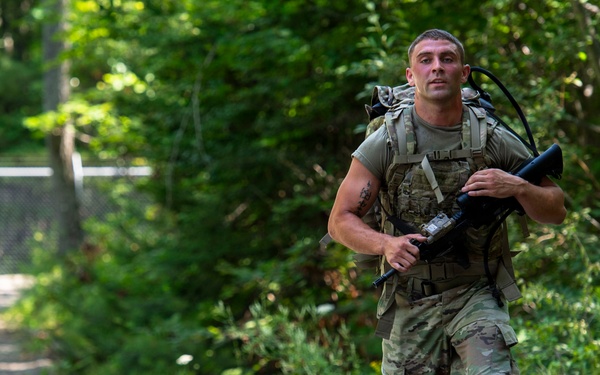Two military personnel in full combat gear are moving along a gravel path surrounded by dense green vegetation. The foreground soldier, wearing OCP (Operational Camouflage Pattern) uniform, carries a rifle and a loaded rucksack, suggesting a ruck march or field training exercise. A second soldier is visible in the background following the same path.