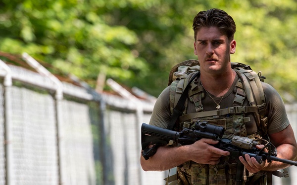 A male soldier in full military gear is walking along a path flanked by a chain-link fence and green trees. He is wearing OCP (Operational Camouflage Pattern) uniform, a plate carrier vest, and a large rucksack, while carrying what appears to be an M4/M16-style rifle. The image appears to be a professional military photograph taken in a training or base environment.