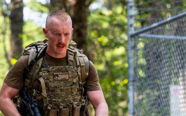 A male soldier in full tactical gear is moving at a jogging pace along a path bordered by trees and a chain-link fence. He is wearing MultiCam camouflage uniform and plate carrier vest, carrying a rifle and a rucksack. The setting appears to be a military training or competition event.