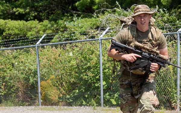 A soldier in full combat gear is running along a gravel path next to a chain-link fence topped with barbed wire. He is wearing MultiCam/OCP camouflage uniform, a boonie hat, tactical vest, and carrying what appears to be an M4/M16-style assault rifle. The setting appears to be a military training facility or base perimeter during daytime.